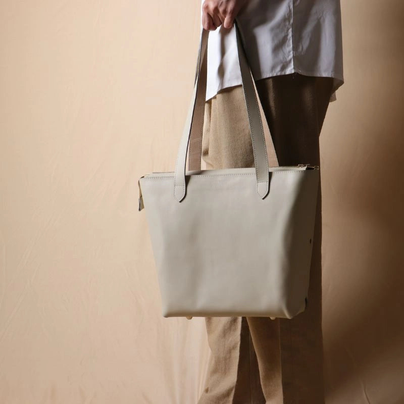 Woman wearing light brown trousers, holding a white leather tote bag with zipper in her hand