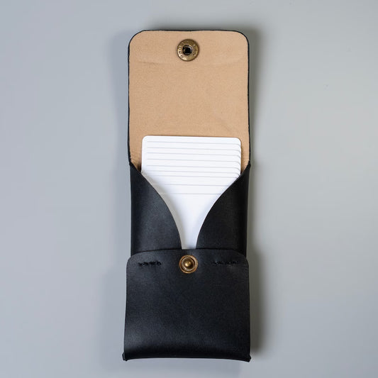 A black full grain leather card holder or card wallet with a beige tan felt lining on the interior flap and brass snap button, shown open with white palstic cards partially inserted, on a light gray background.
