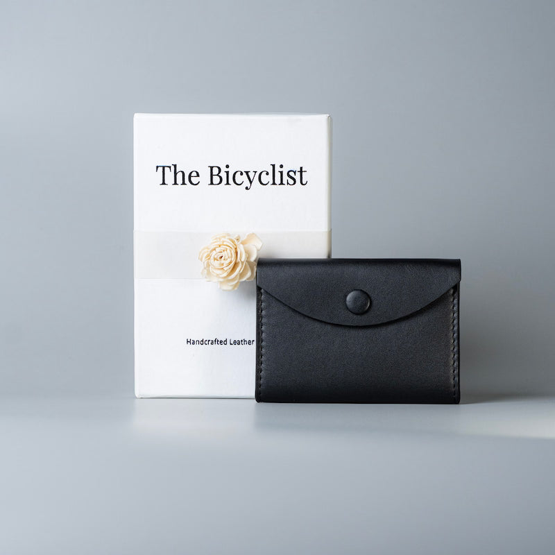 A black full grain leather card holder wallet with a snap-button flap stands in front of a white product box labeled 'The Bicyclist' on a light gray background.
