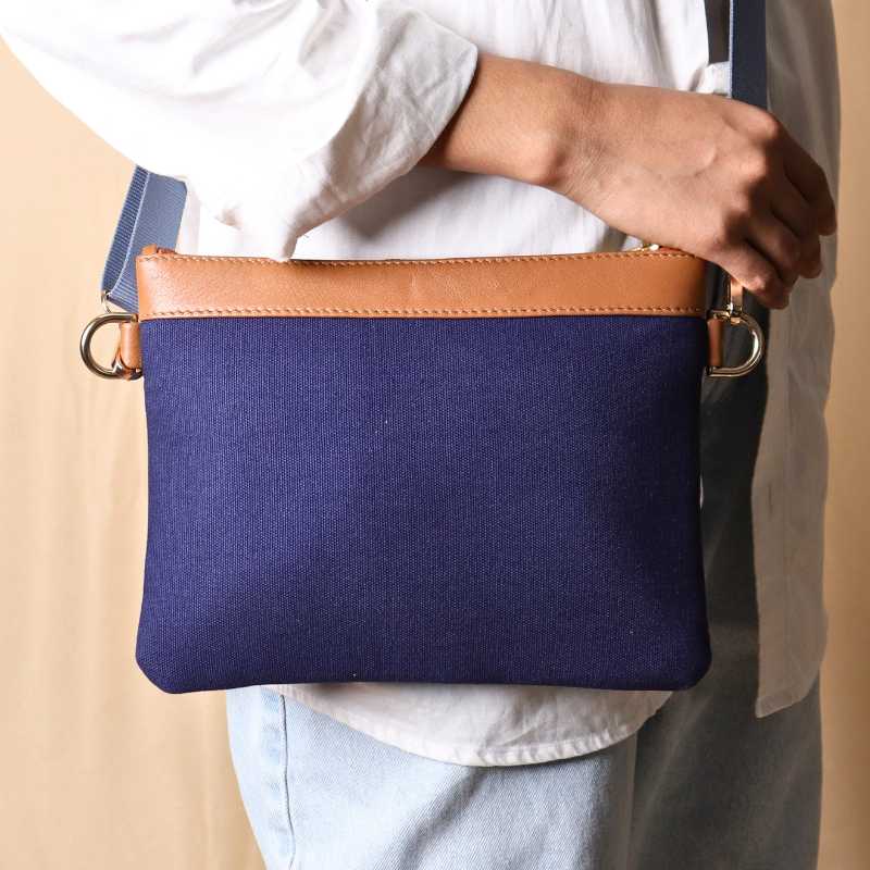 Woman in a white shirt and light blue denims carrying over her shoulder, a deep blue canvas crossbody sling bag with tan leather trims on the top and blue nylon strap
