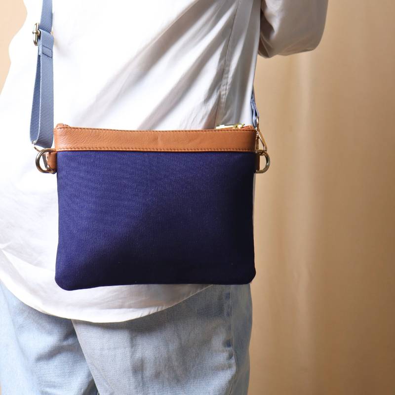 Woman in a white shirt and light blue denims carrying over her shoulder, a deep blue canvas crossbody sling bag with tan leather trims on the top and blue nylon strap