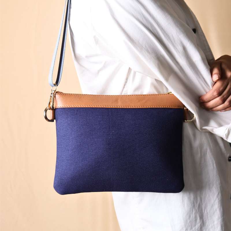 Woman in a white shirt and light blue denims carrying over her shoulder, a deep blue canvas crossbody sling bag with tan leather trims on the top and blue nylon strap