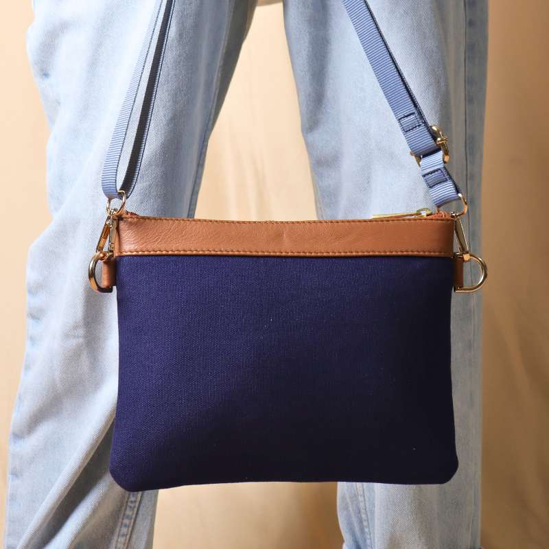 Woman in a white shirt and light blue denims carrying on her hand, a deep blue canvas crossbody sling bag with tan leather trims on the top and blue nylon strap.