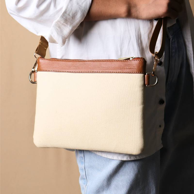 Woman in a white shirt and light blue denims carrying over her shoulder, a Off white canvas crossbody sling bag with tan leather trims on the top and tan nylon strap.