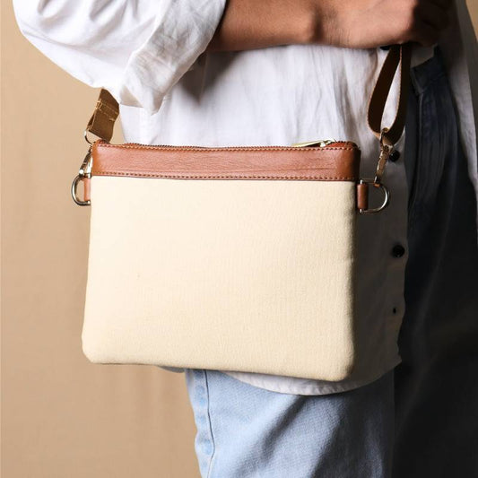 Woman in a white shirt and light blue denims carrying over her shoulder, a Off white canvas crossbody sling bag with tan leather trims on the top and tan nylon strap.