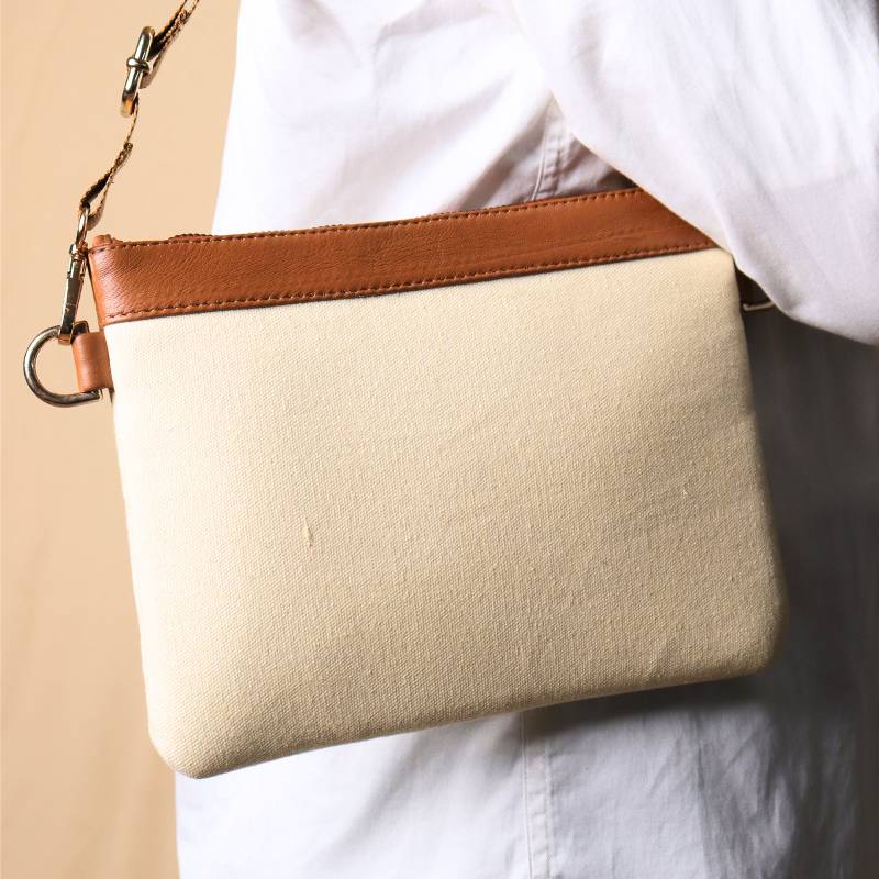 Woman in a white shirt and light blue denims carrying over her shoulder, a Off white canvas crossbody sling bag with tan leather trims on the top and tan nylon strap.