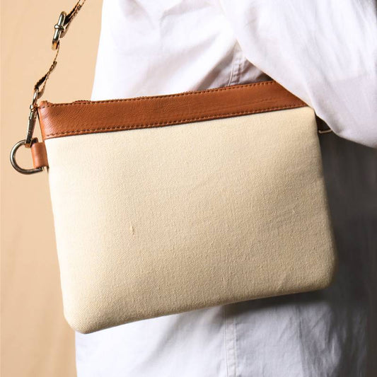 Woman in a white shirt and light blue denims carrying over her shoulder, a Off white canvas crossbody sling bag with tan leather trims on the top and tan nylon strap.