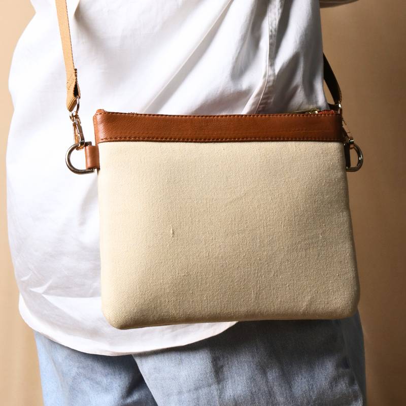 Woman in a white shirt carrying over her shoulder, a Off white canvas crossbody sling bag with tan leather trims on the top and tan nylon strap.