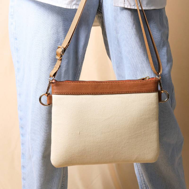 Woman in a white shirt and light blue denims carrying in her hand, a Off white canvas crossbody sling bag with tan leather trims on the top and tan nylon strap.