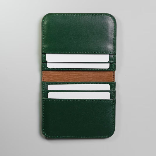 A dark green full grain leather card holder wallet spread open with a tan brown sheep leather lining in the center, with white plastic cards inserted in the card slots made by, 'The Bicyclist' kept flat on a light gray background.