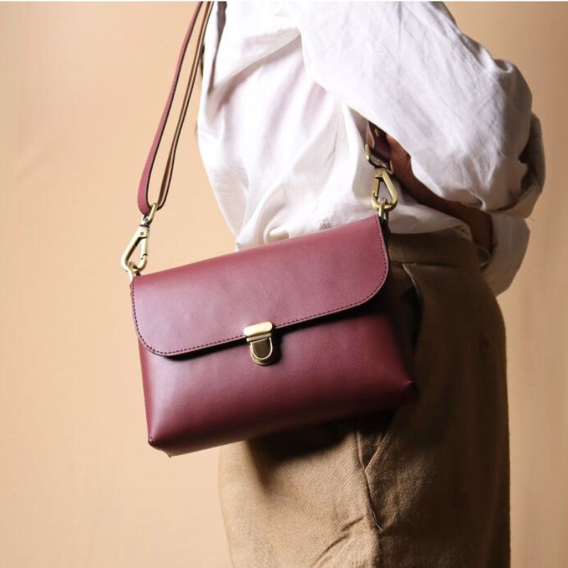 Woman in a white shirt carrying over her shoulder a maroon leather crossbody shoulder side bag with a flap and a light gold push lock