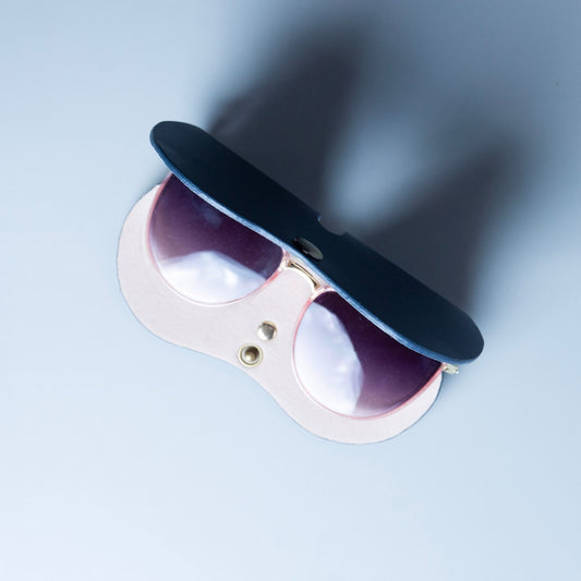 Top view image of a navy blue full grain leather eyeglass sleeves made by 'The Bicyclist' that is open and shows a beige felt lining on the inside along with a pair of sunglasses facing up and snap button on a soft grey background.
