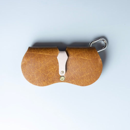 Back view of a tan textured leather spectacle case from 'The Bicyclist' with a silver metal carabiner clip and a central silver metal accent, photographed from directly above on a light blue background.