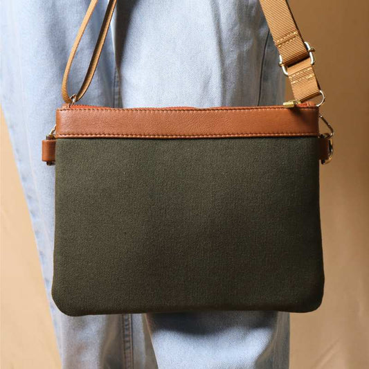 Woman in light blue denims carrying a dark green canvas crossbody sling bag with tan leather trims on the top and green nylon strap.