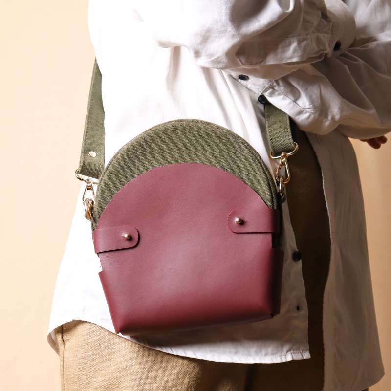 Woman in beige pants carrying a dark green canvas crossbody sling in a maroon leather case across her shoulder as a crossbody