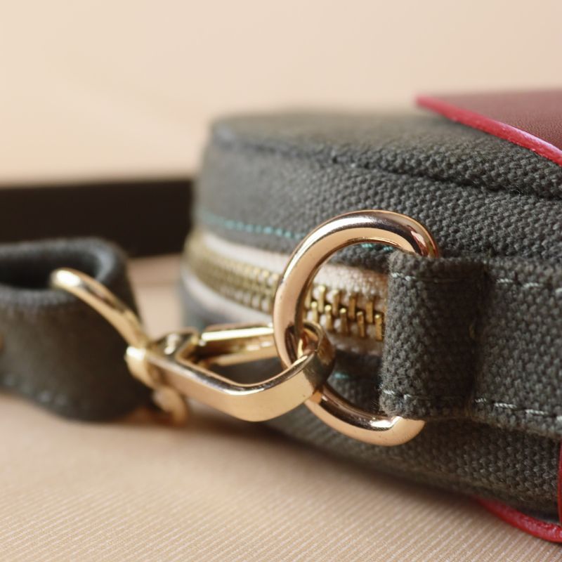 Detailed Gold Metal Fitting view of a Dark Green Canvas Crossbody sling bag in a maroon leather case with its canvas strap extended