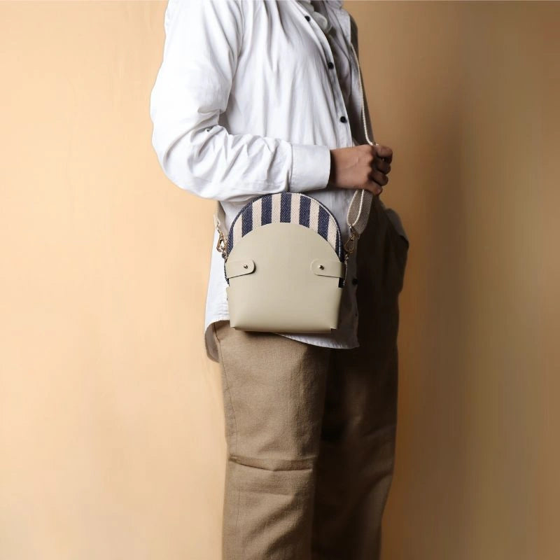 Woman in white shirt and beige pants carrying around her shoulder a canvas bag with blue and white stripe weave inside a off-white leather case