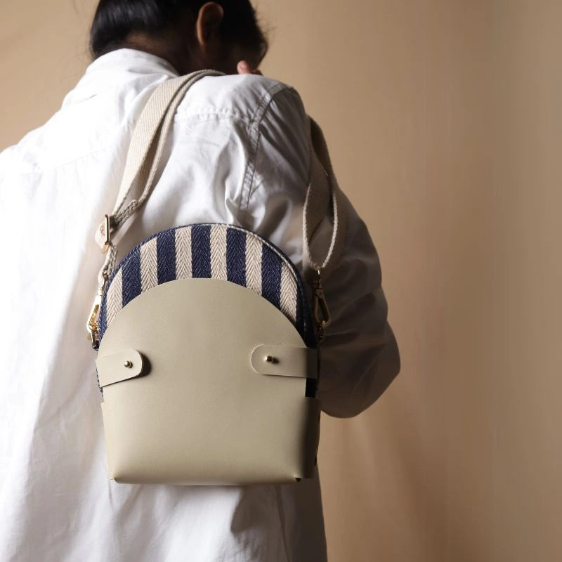 Back View of a Woman in white shirt and beige pants carrying over her shoulder a canvas bag with blue and white stripe weave inside a off-white leather case