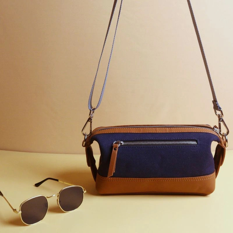Front view of a navy blue cotton canvas and tan leather dopp kit with a zip, with extended straps and a pair of sunglasses