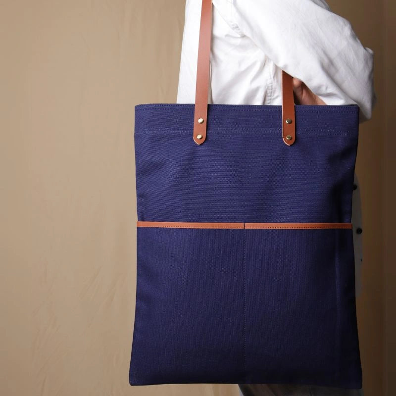 Woman in a white shirt carrying over her shoulder, a dark blue canvas tote with leather straps and leather trim front pockets
