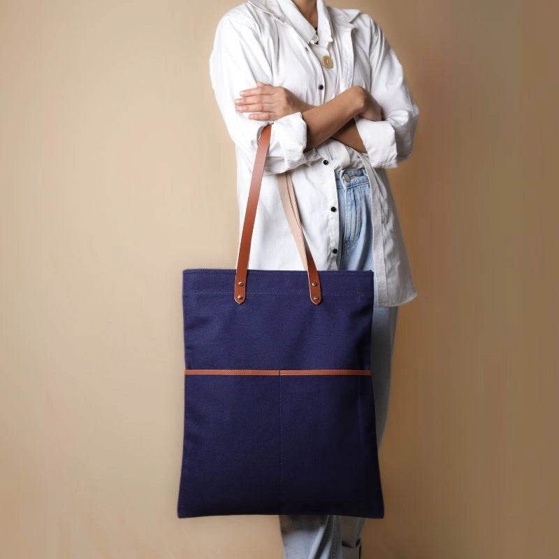 Woman in a white shirt and light blue denims holding in her hand, a dark blue canvas tote with leather straps and leather trim front pockets