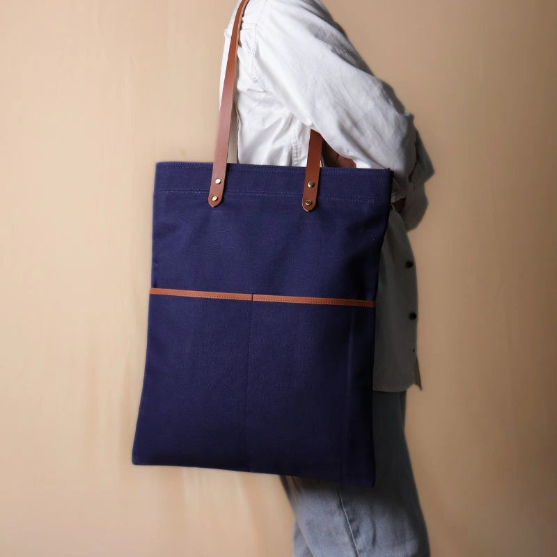 Woman in a white shirt and light blue denims holding in her hand, a dark blue canvas tote with leather straps and leather trim front pockets