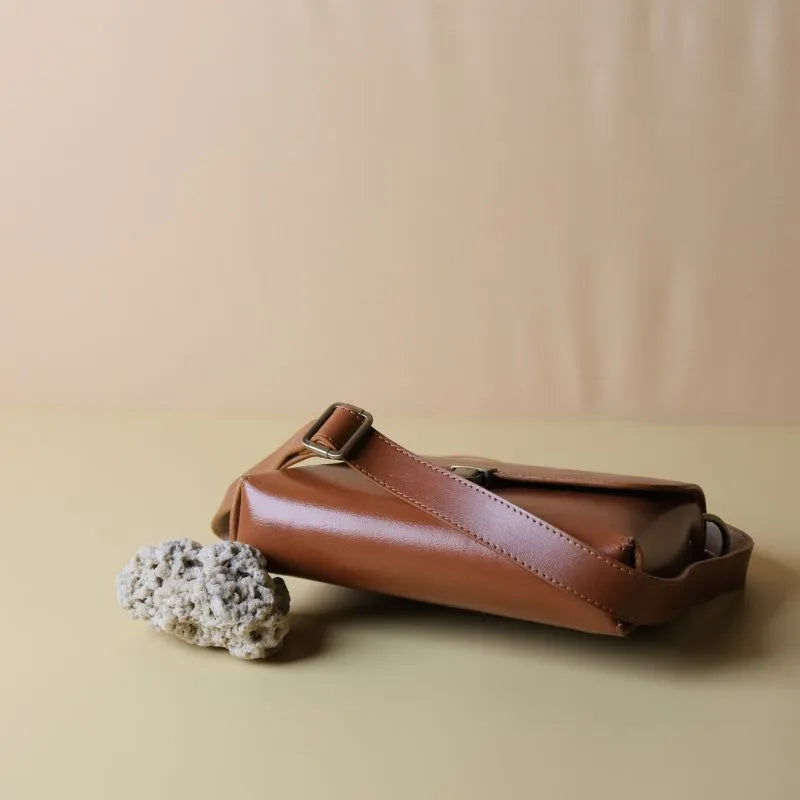 Bottom Image of a Cognac Tan  Leather Crossbody sling bag with straps and a flap with a light gold push lock raised with the support of a white coral on the left bottom corner on a light peach background