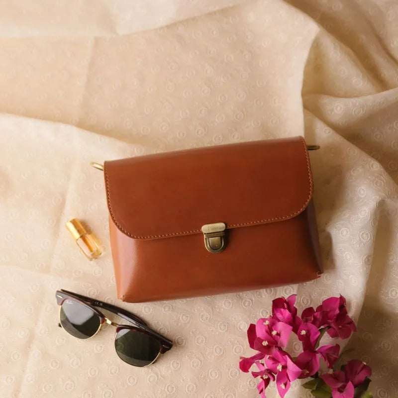 Cognac Tan Leather Crossbody sling bag with a flap, a light gold push lock and a pair of dark glasses, pink bougainvillea flowers are kept next to it