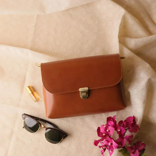 Cognac Tan  Leather Crossbody sling bag with  a flap, a light gold push lock and a pair of dark glasses, pink bougainvillea flowers are kept next to it