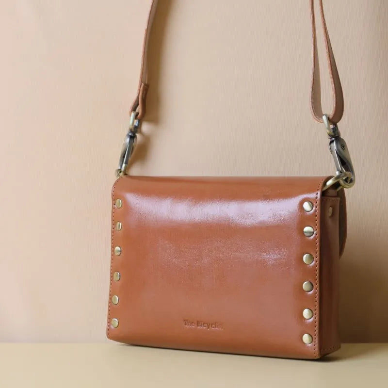 Cognac Tan Leather Crossbody sling bag with straps and a flap with a light gold fittings on a peach background
