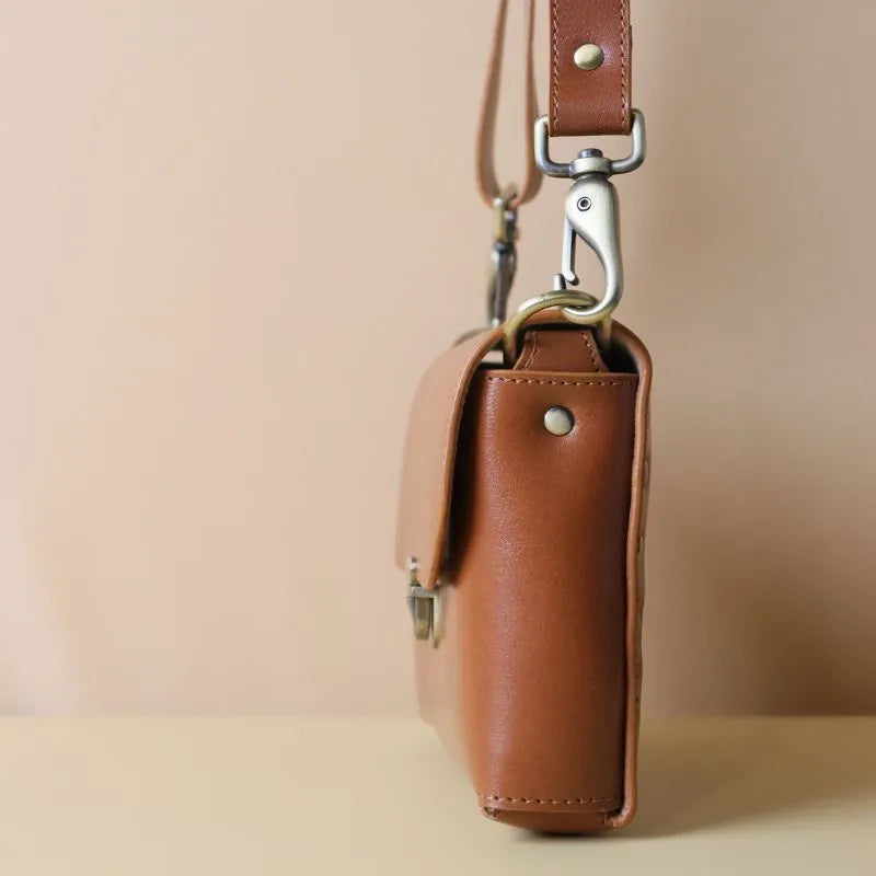 Side View of a Cognac Tan Leather Crossbody sling bag with straps and a flap with a light gold strap fittings on a peach background