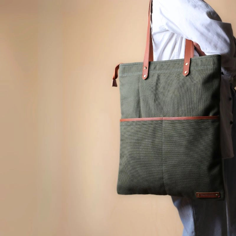 Woman in a white shirt and light blue denims carrying over her shoulder, a dark green canvas tote with leather straps and leather trim front pocket