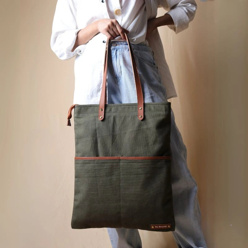 Woman in a white shirt and light blue denims carrying on her hand, a dark green canvas tote with leather straps and leather trim front pocket