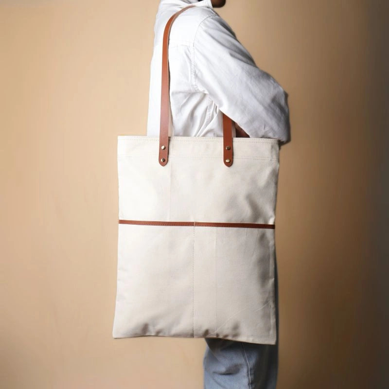 Woman in a white shirt and light blue denims carrying over her shoulder, a off-white canvas tote with leather straps and leather trim front pocket