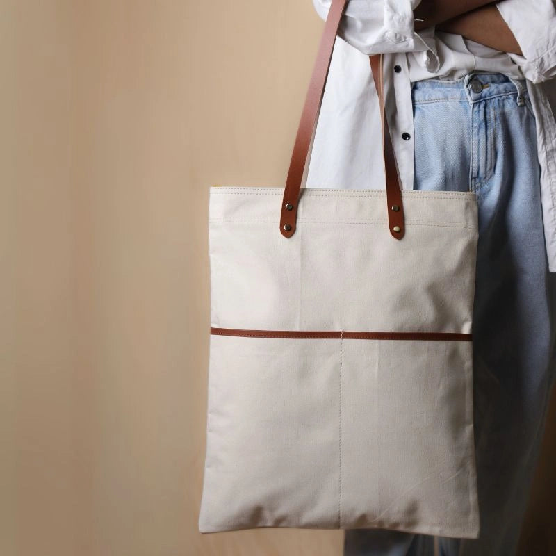 Woman in a white shirt and light blue denims holding in her hand, a off-white canvas tote with leather straps and leather trim front pocket