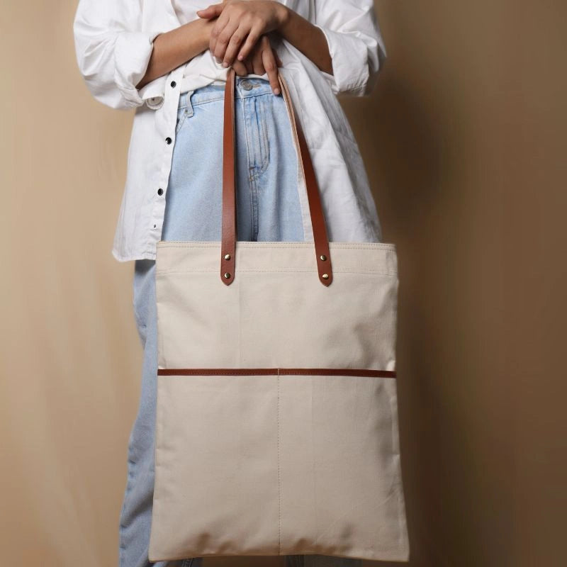 Woman in a white shirt and light blue denims carrying in her hand, a off-white canvas tote with leather straps and leather trim front pocket