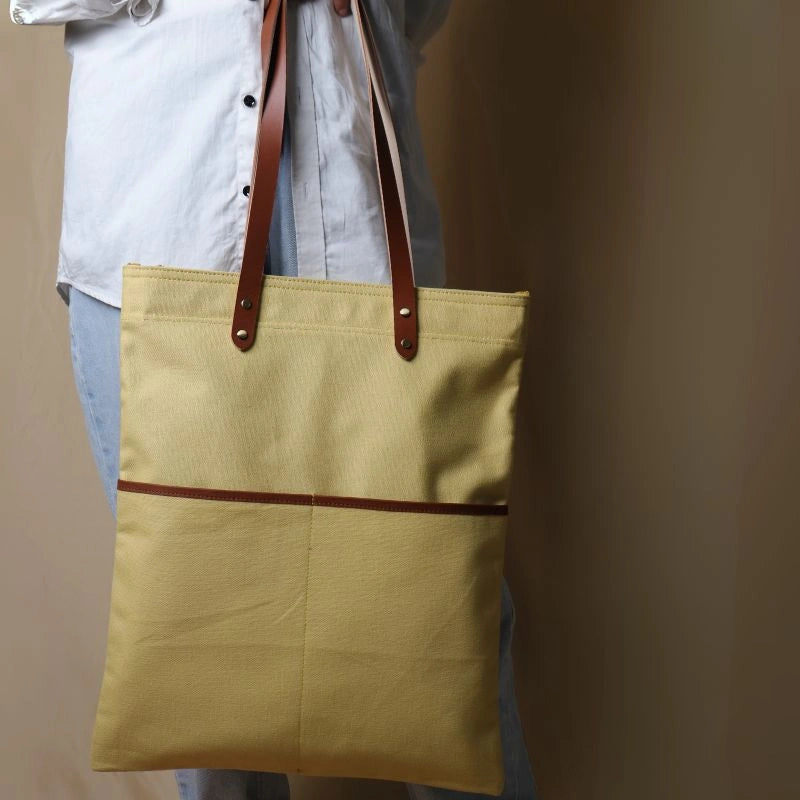 Woman in a white shirt and light blue denims holding in her hand, a light yellow canvas tote with leather straps and leather trim front pocket
