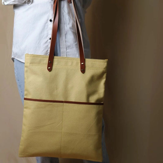 Woman in a white shirt and light blue denims holding in her hand, a light yellow canvas tote with leather straps and leather trim front pocket