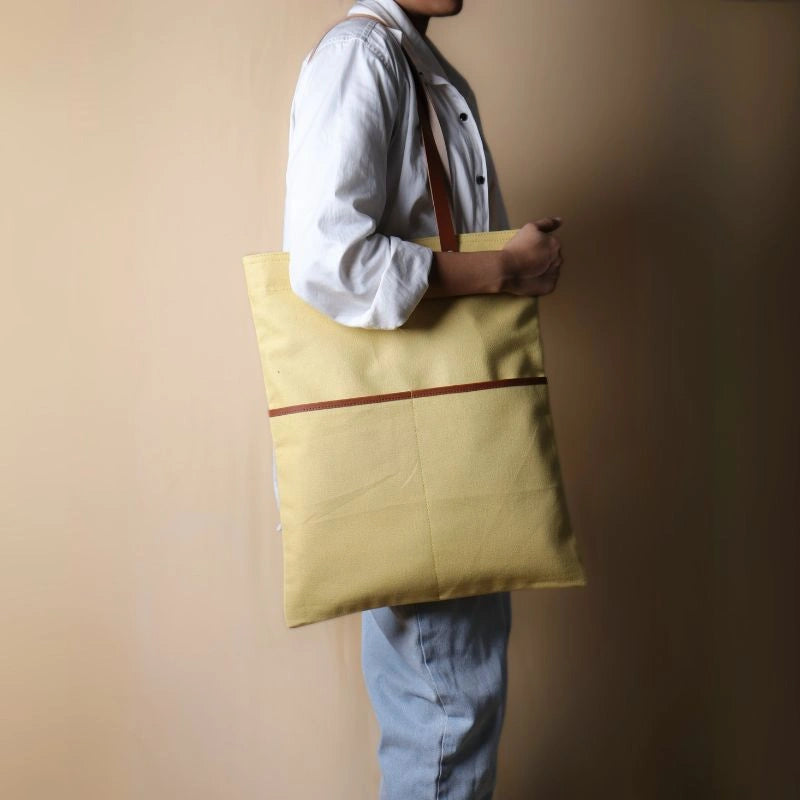 Woman in a white shirt and light blue denims carrying over her shoulder, a light yellow canvas tote with leather straps and leather trim front pocket
