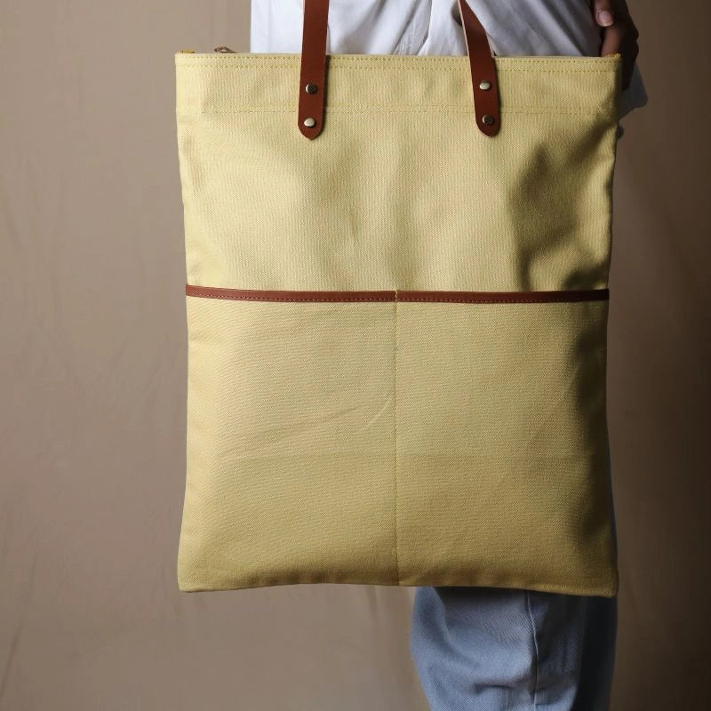 Woman in a white shirt and light blue denims carrying over her shoulder, a light yellow canvas tote with leather straps and leather trim front pocket