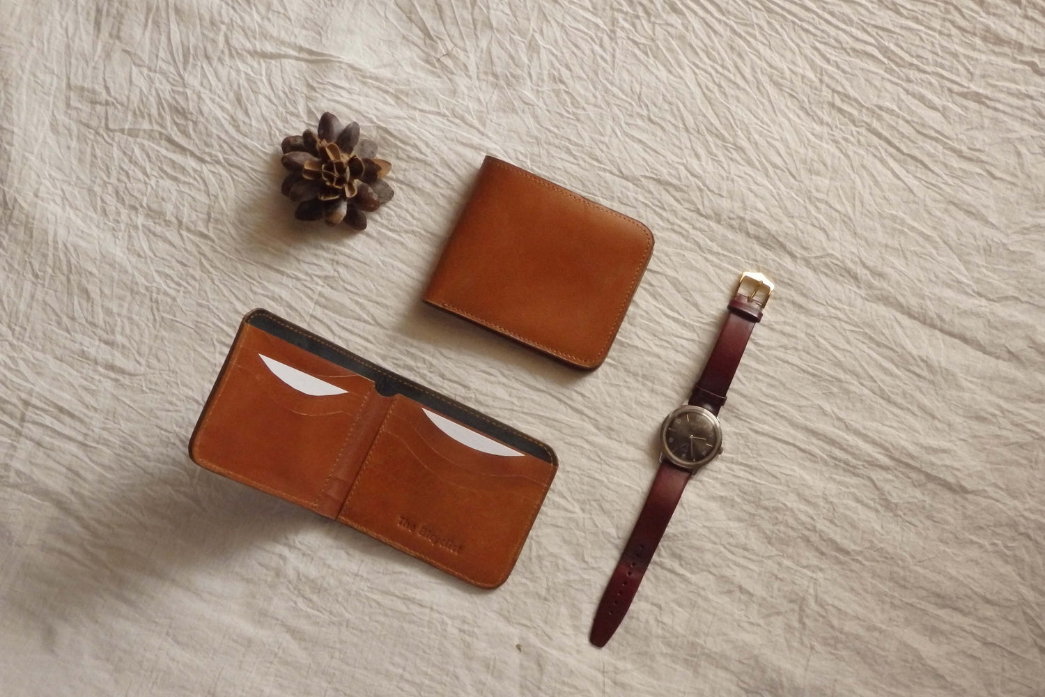 Handmade Veg Tanned Full Grain Bovine Leather Bifold Card Wallet in Tan with an analogue wrist watch and a pine cone