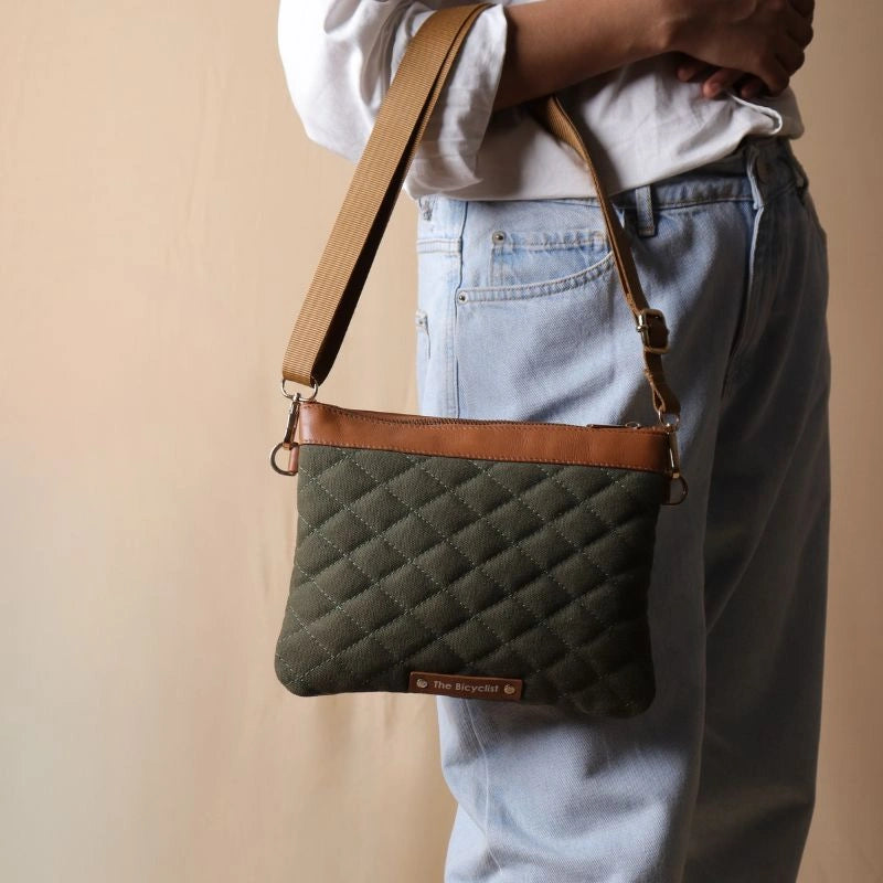 Woman in white shirt and light blue denims carrying a dark green quilted canvas sling bag with tan leather trim in her hand