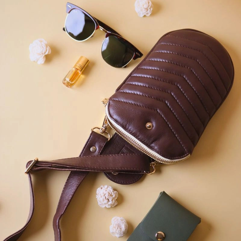 A Dark Brown Quilted Leather Pouch Bag with a golden zip and a card case, sunglasses, perfume kept next to it with white flowers strewn around