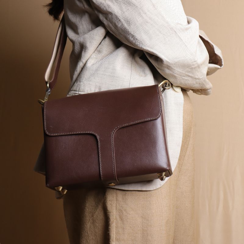 Close up view of Woman in an off-white blazer carrying a dark brown leather crossbody sling bag with flap around the shoulder