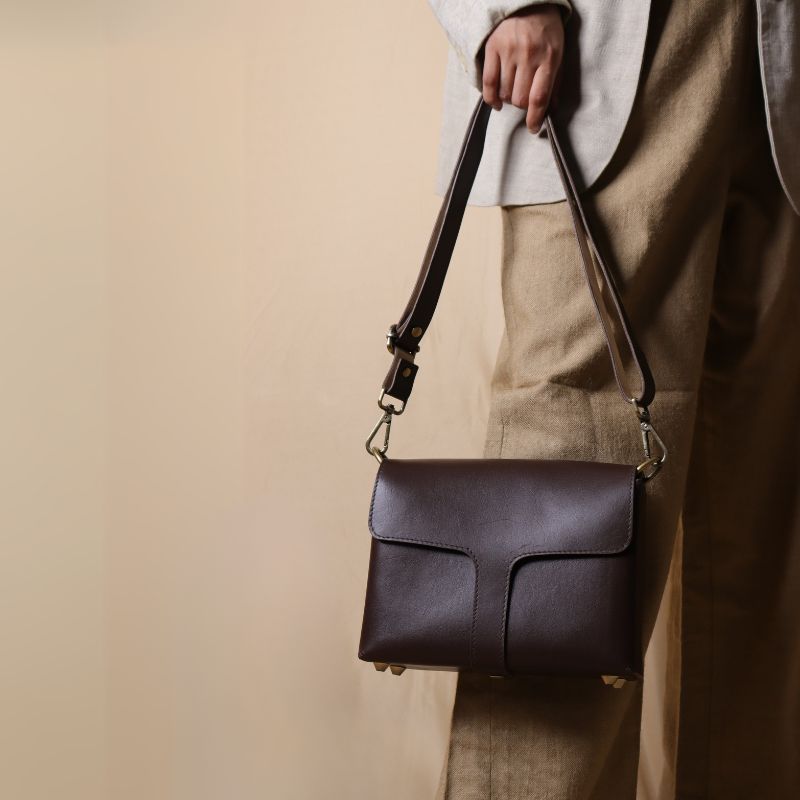 Woman in an dark beige trousers carrying a dark brown leather crossbody sling bag with flap around the shoulder