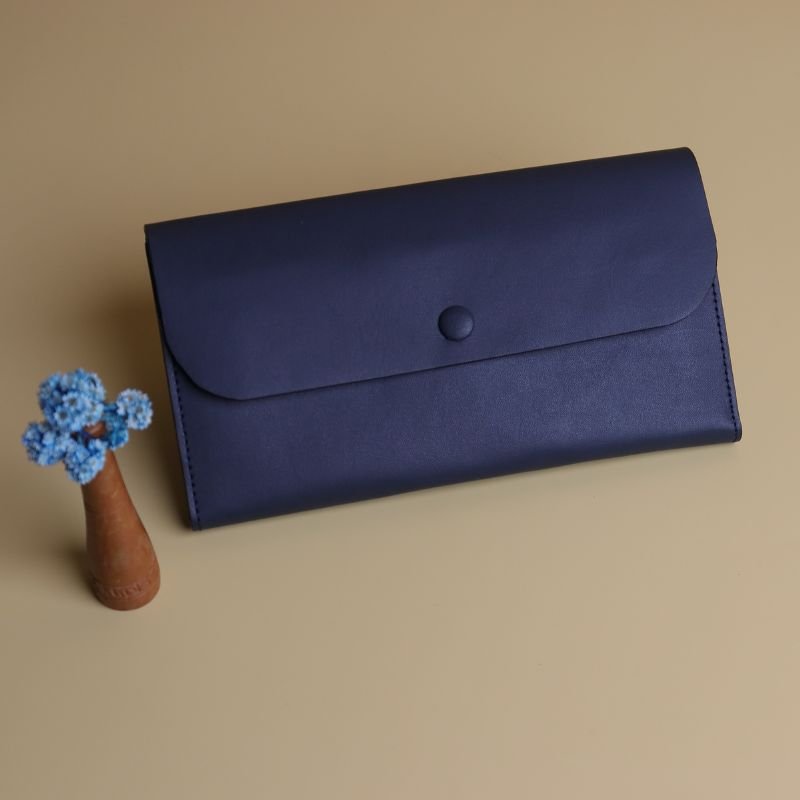 Front View of a Large Long Wallet for Women Handmade in Deep Blue Leather with a Flap and a Button covered with leather and a small flower vase with light blue dry flowers