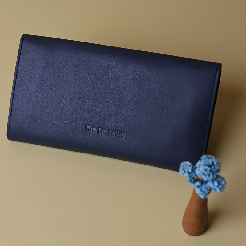 Back View of a Large Long Wallet for Women Handmade in Deep Blue leather with the name of the brand embossed in the lower center along with a tiny vase with light blue dry flowers on a cream background