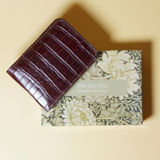 Maroon crocodile texture leather wallet on a floral-patterned box with 'The Bicyclist' branding.
