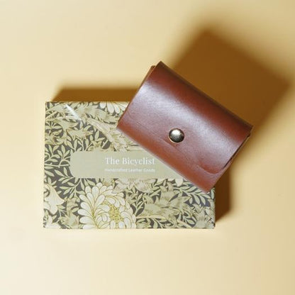 Tan Brown leather wallet on a floral-patterned box with 'The Bicyclist' branding on a beige background