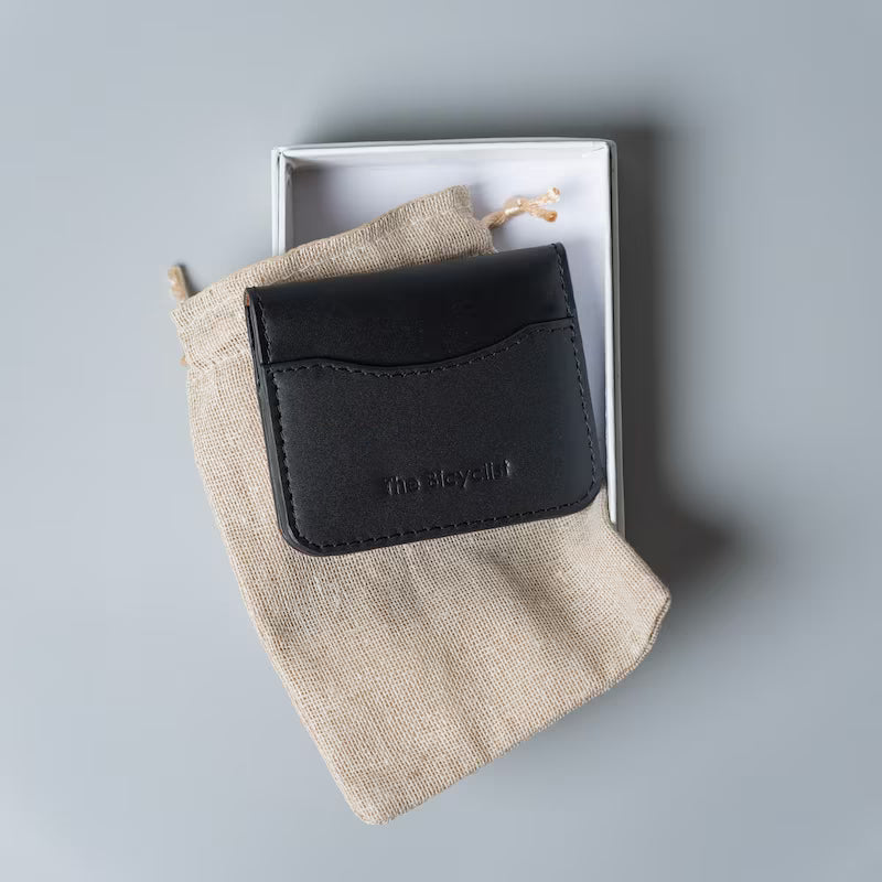 A black leather card holder wallet with debossed 'The Bicyclist' branding, resting on a beige linen drawstring pouch inside an open white box, set against a solid gray background.